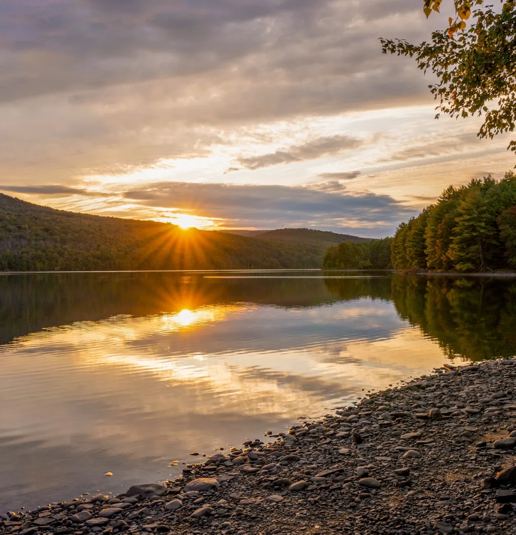 Sunset over a Catskills reservoir, with the sun low behind wooded hills and a stony shoreline in the foreground